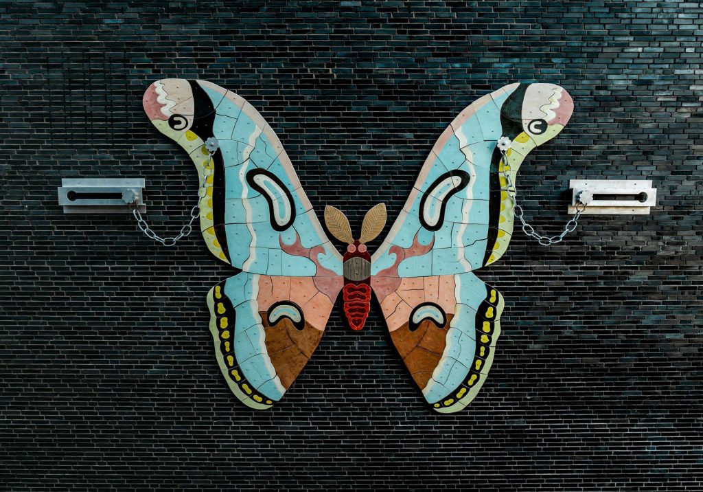 Farbige Schmetterlingsskulptur mit Ketten an den Flügelspitzen, befestigt an einer dunklen Ziegelwand.