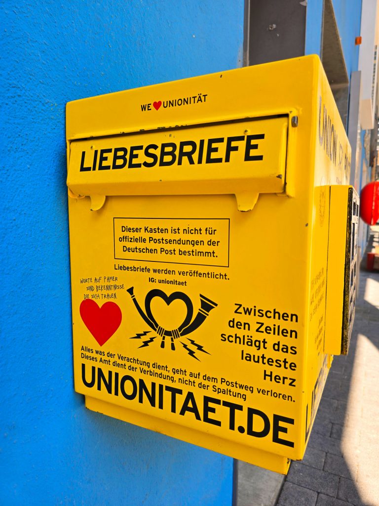 Gelber Briefkasten mit der Aufschrift „Liebesbriefe“ und „unionitaet.de“ an blauer Wand, gestaltet als Symbol für Verbindung und Ausdruck.