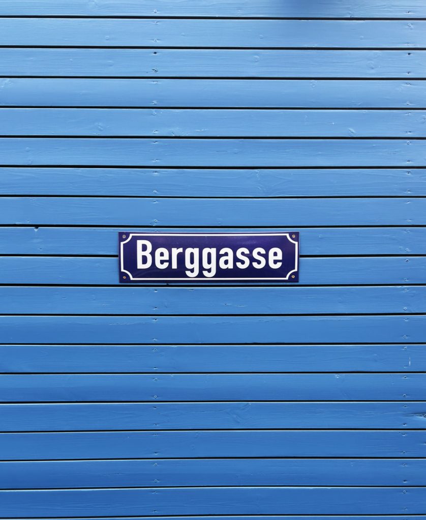 Straßenschild „Berggasse“ auf blauer Holzverkleidung mit waagerechter Lattenstruktur.