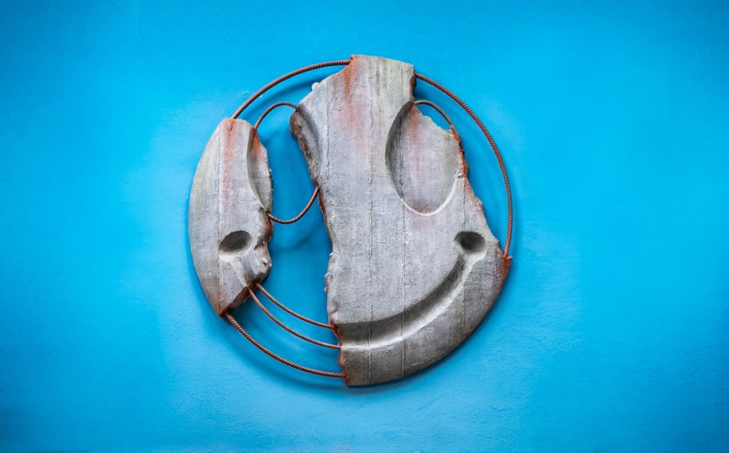 Betonrelief in Form eines zerbrochenen Smileys mit rostigem Bewehrungsstahl, montiert auf blauer Wand.