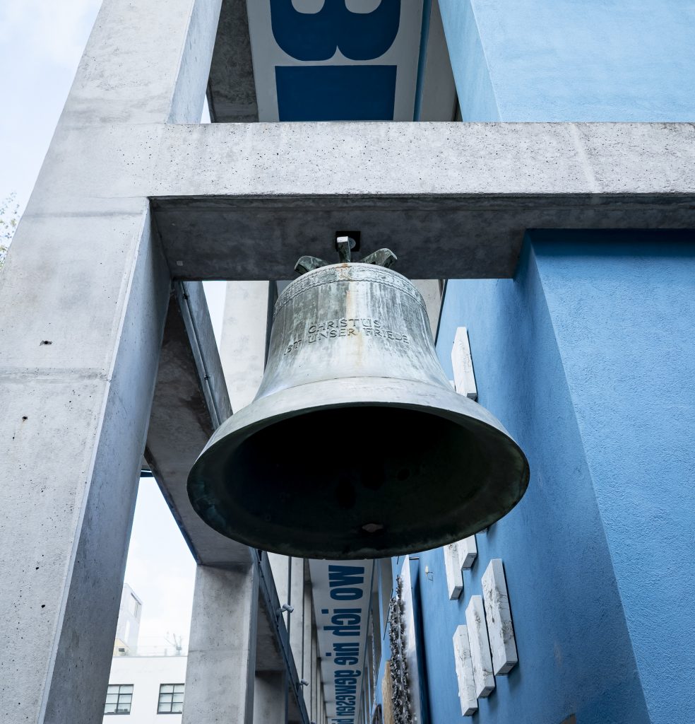 Glocke mit der Inschrift „Christus ist unser Friede“, frontal von unten vor Beton und blauer Fassade.