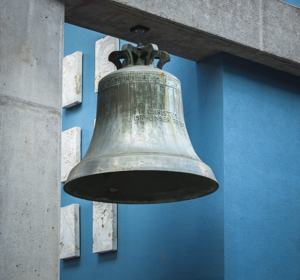 Glocke mit der Inschrift „Christus ist unser Friede“, frontal von unten vor Beton und blauer Fassade.