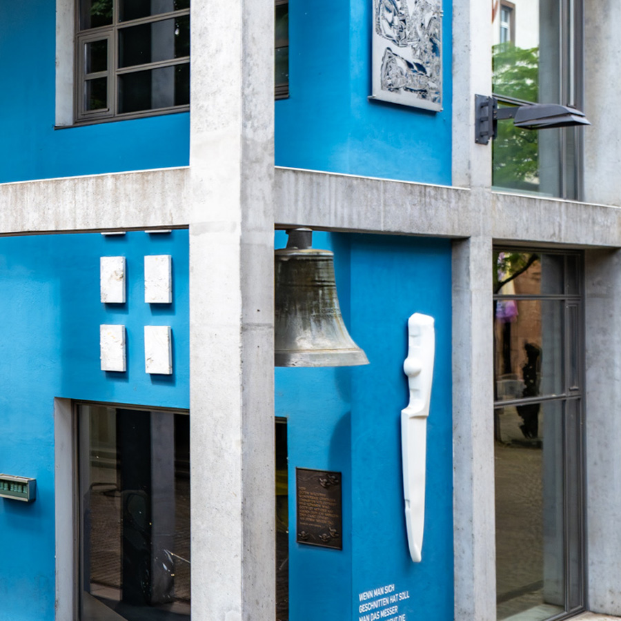 Seitlicher Blick auf Bronzeglocke und weißes Messerrelief an blauer Fassade, umgeben von Kunstwerken.