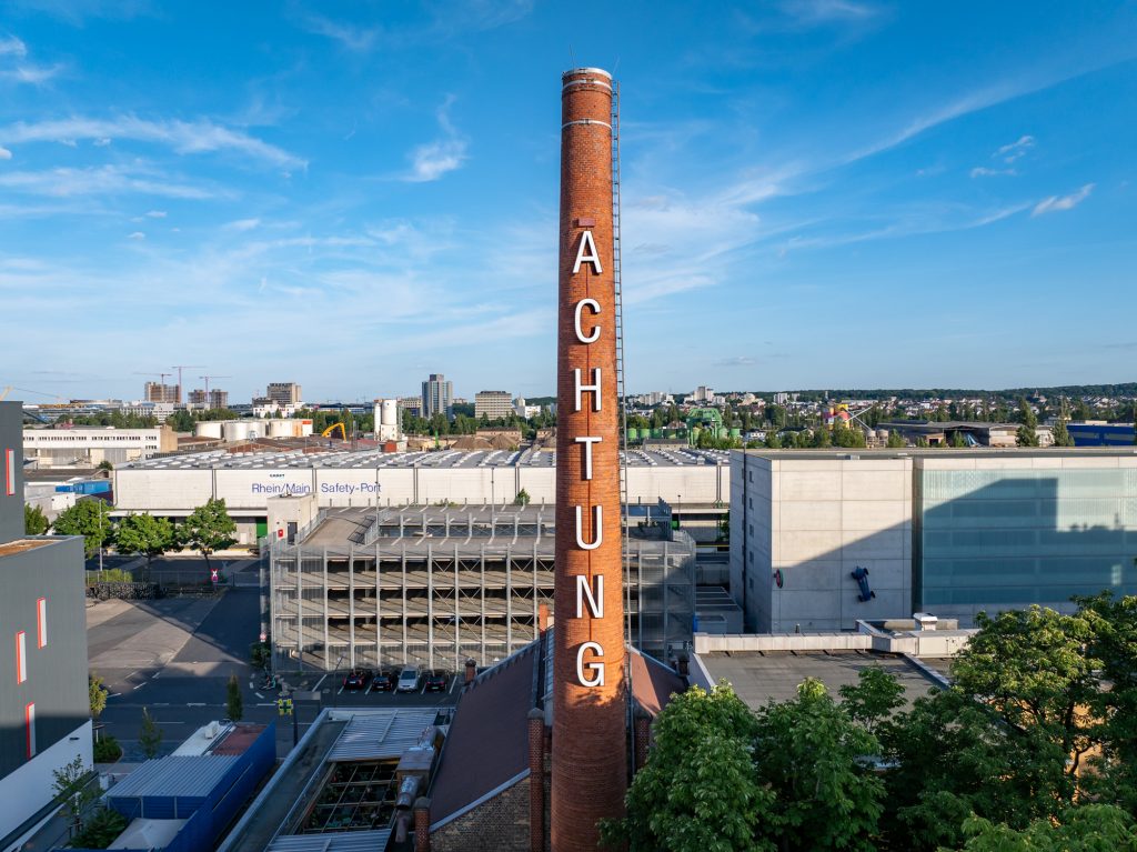 Ziegelroter Schornstein mit großem weißen Schriftzug „ACHTUNG“, im Hintergrund Industriegebiet und blauer Himmel.