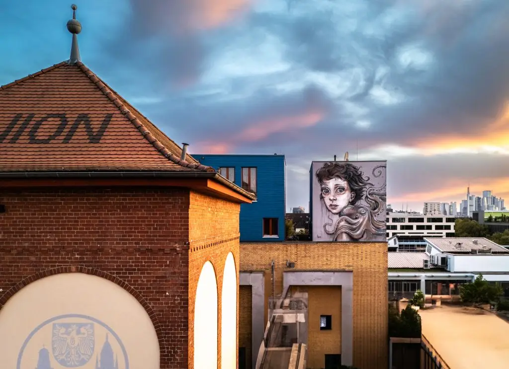 Turm mit UNION-Schriftzug, daneben bemalte Wand mit großem Frauenporträt auf Hausdach bei Sonnenuntergang.
