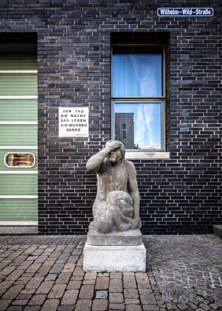 Steinskulptur einer knienden Figur mit der Hand auf der Stirn, vor schwarzer Klinkerwand und Texttafel „DER TAG …“.