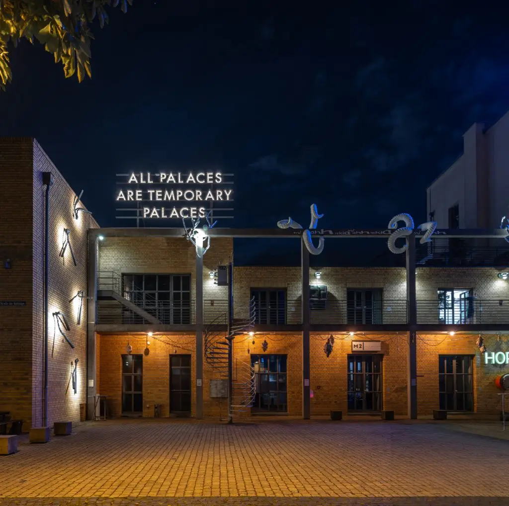 Panorama mit leuchtenden Fassaden, Kunstinstallationen und dem Schriftzug „ALL PALACES ARE TEMPORARY PALACES“.