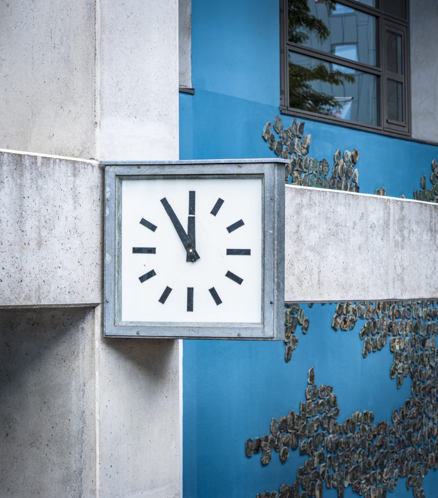 Uhr an Betonträger vor blauer Wand mit Kunstinstallation, Zeiger stehen auf fünf vor zwölf.