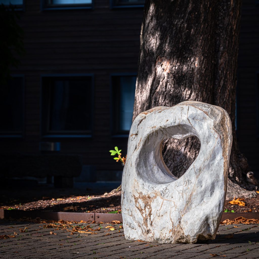 Herzförmige Öffnung in hellem, marmorähnlichem Steinblock, platziert auf gepflastertem Boden im Schatten eines Baums mit Hintergrundstruktur und Herbstlaub.