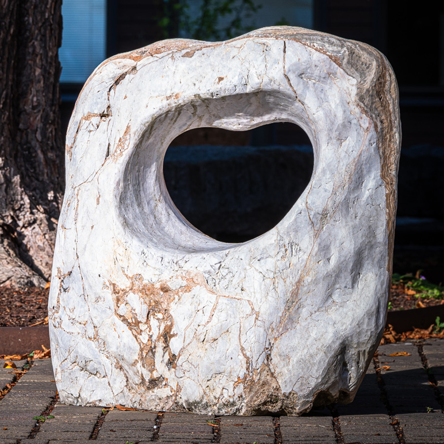 Herzförmig durchbrochener heller Stein mit glatter Oberfläche, auf Pflaster zwischen Bäumen und Gebäudestruktur platziert.