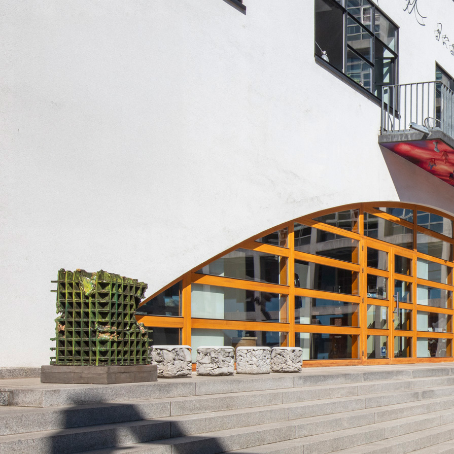 Vier helle Sitzquader und ein grünes, rasterartiges Kunstobjekt auf Stufen vor Fensterfassade mit gelbem Holzrahmen.