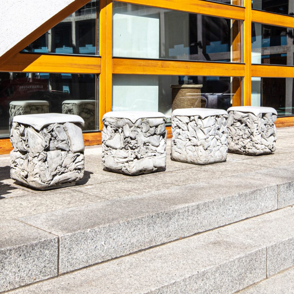 Vier helle Quader mit rauer, gepresster Oberfläche und glatter Oberseite auf Steinstufen vor Glasfassade.