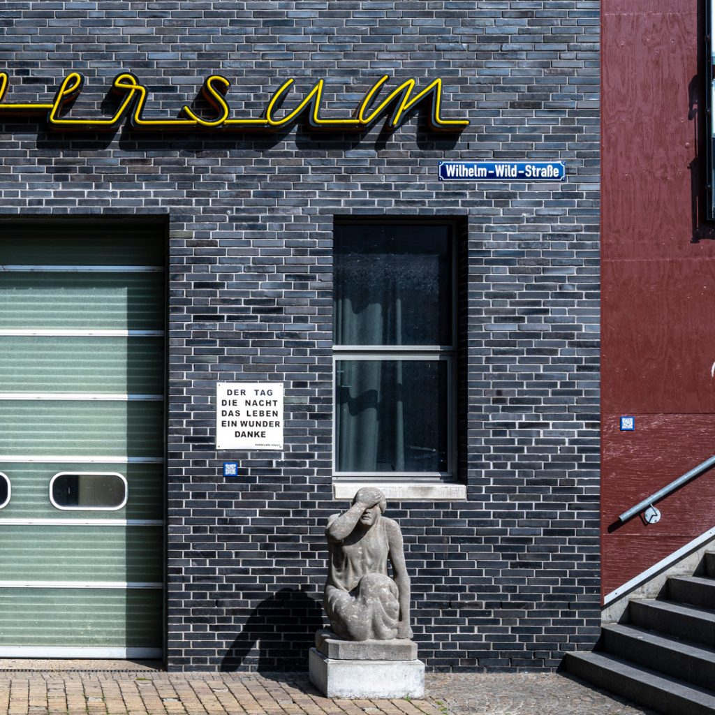 Skulptur einer sitzenden Figur mit erhobener Hand vor dem Gesicht, platziert vor dunkler Backsteinwand.