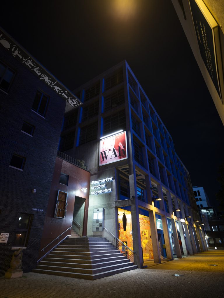 Nachtansicht mit beleuchteter Treppe und Schriftzug „Baumarten sterben angenehm“.