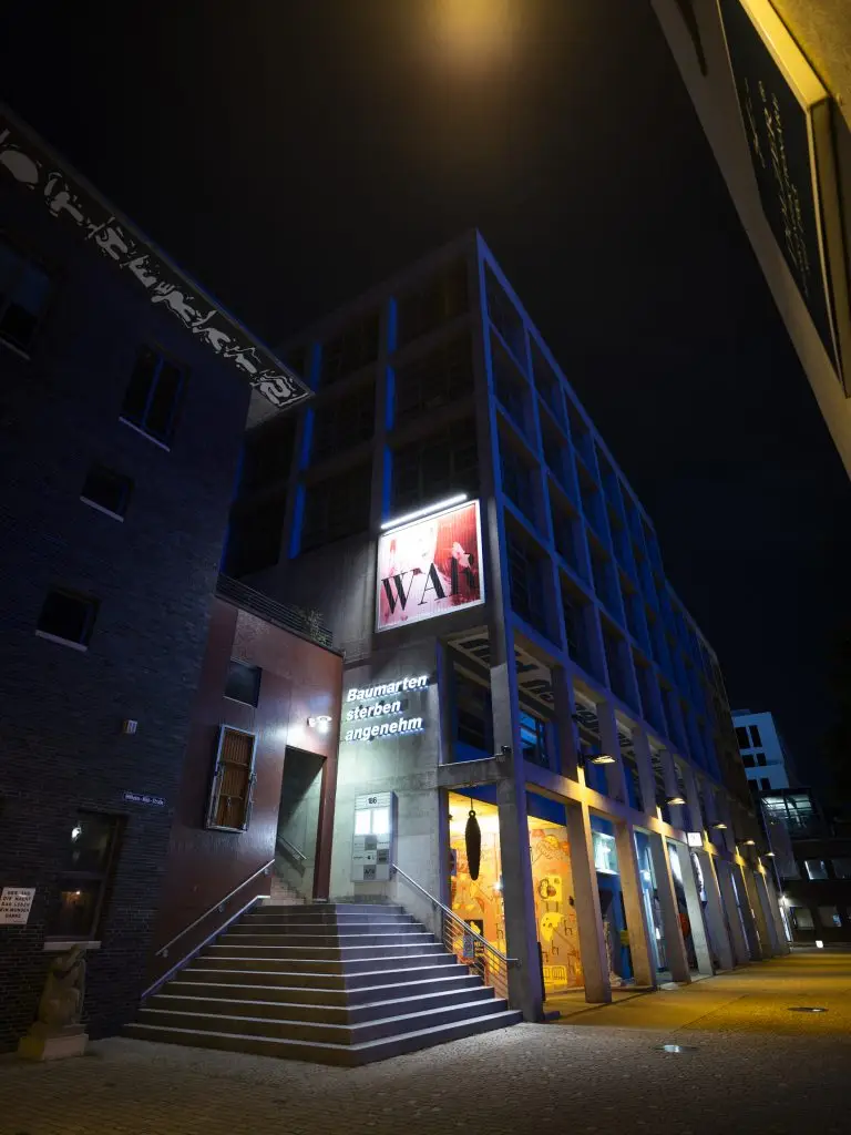 Nachtansicht mit beleuchteter Treppe und Schriftzug „Baumarten sterben angenehm“.