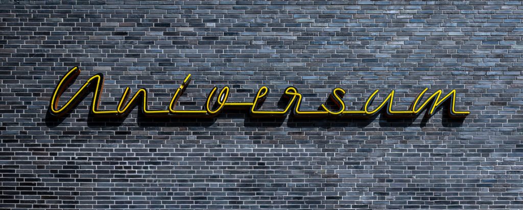 Gelber Neonschriftzug „Universum“ auf dunkler Backsteinwand, frontal aufgenommen.