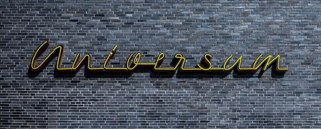 Gelber Neonschriftzug „Universum“ auf dunkler Backsteinwand, frontal aufgenommen.
