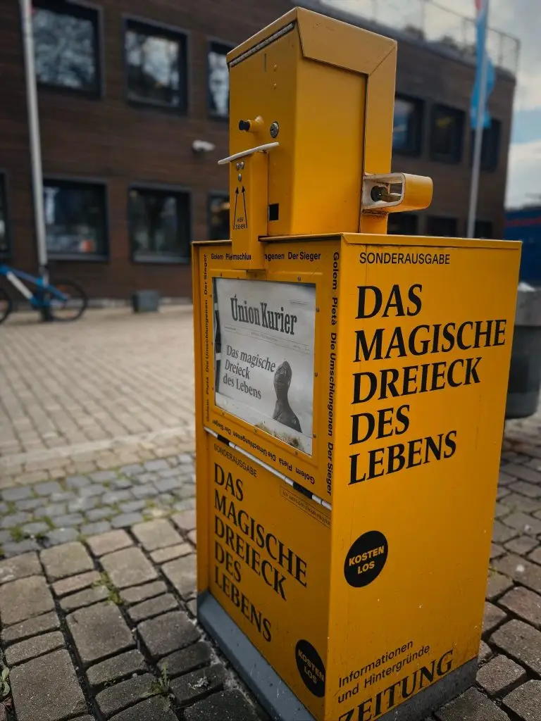 Seitenansicht des gelben Zeitungsautomaten mit dem groß gedruckten Text "Das magische Dreieck des Lebens", Union-Gelände.