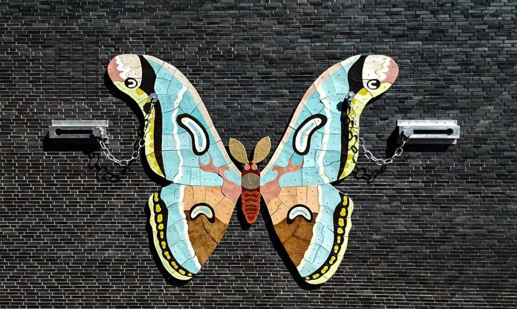 Großes, farbenfrohes Schmetterlings-Wandobjekt, mit Ketten an dunkler Backsteinwand fixiert, Union-Gelände Frankfurt.
