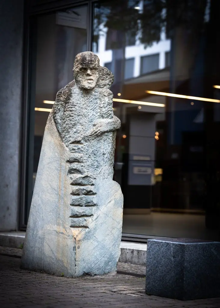 Raue Steinskulptur eines bärtigen Mannes mit markanten Einkerbungen vor Glasfassade, Union-Gelände Frankfurt.