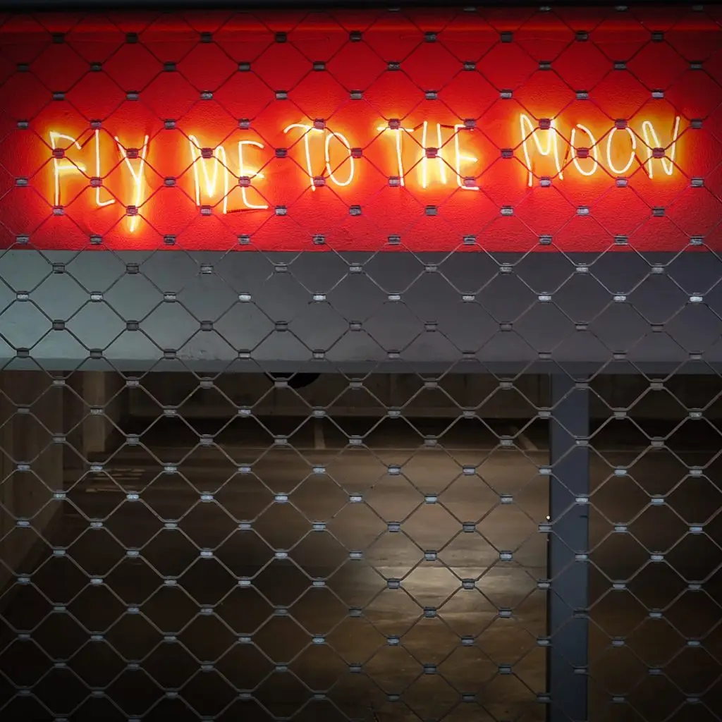 Orangefarbener Neon-Schriftzug "FLY ME TO THE MOON" hinter einem Metallgitter, Union-Gelände Frankfurt.