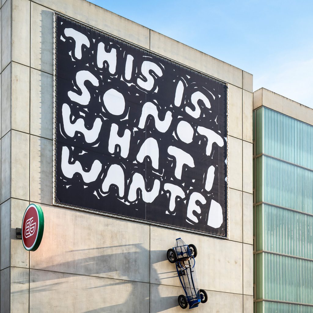 Nahaufnahme des Stefan Marx Kunstwerks: Schwarz-weißes Banner und Sportwagen-Skulptur an Betonwand.