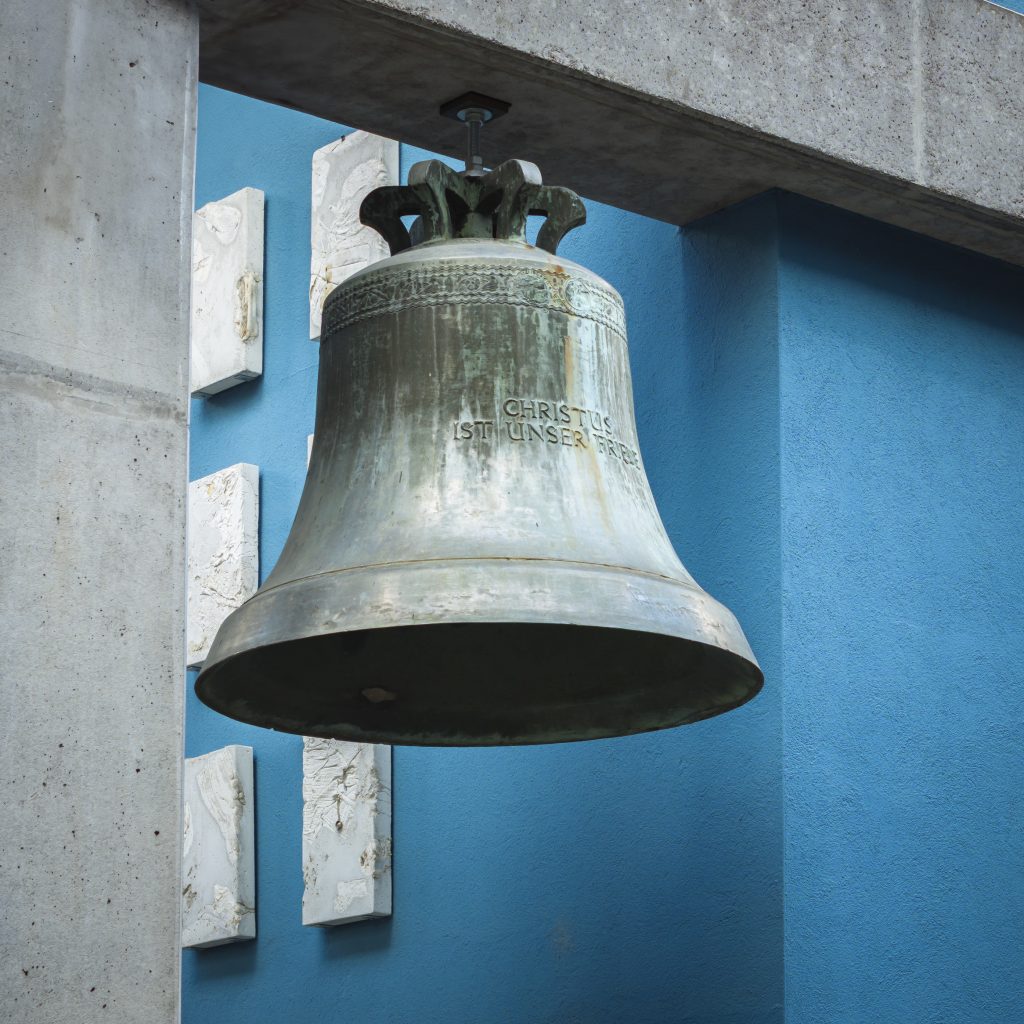 Glocke mit der Inschrift „Christus ist unser Friede“, frontal von unten vor Beton und blauer Fassade.