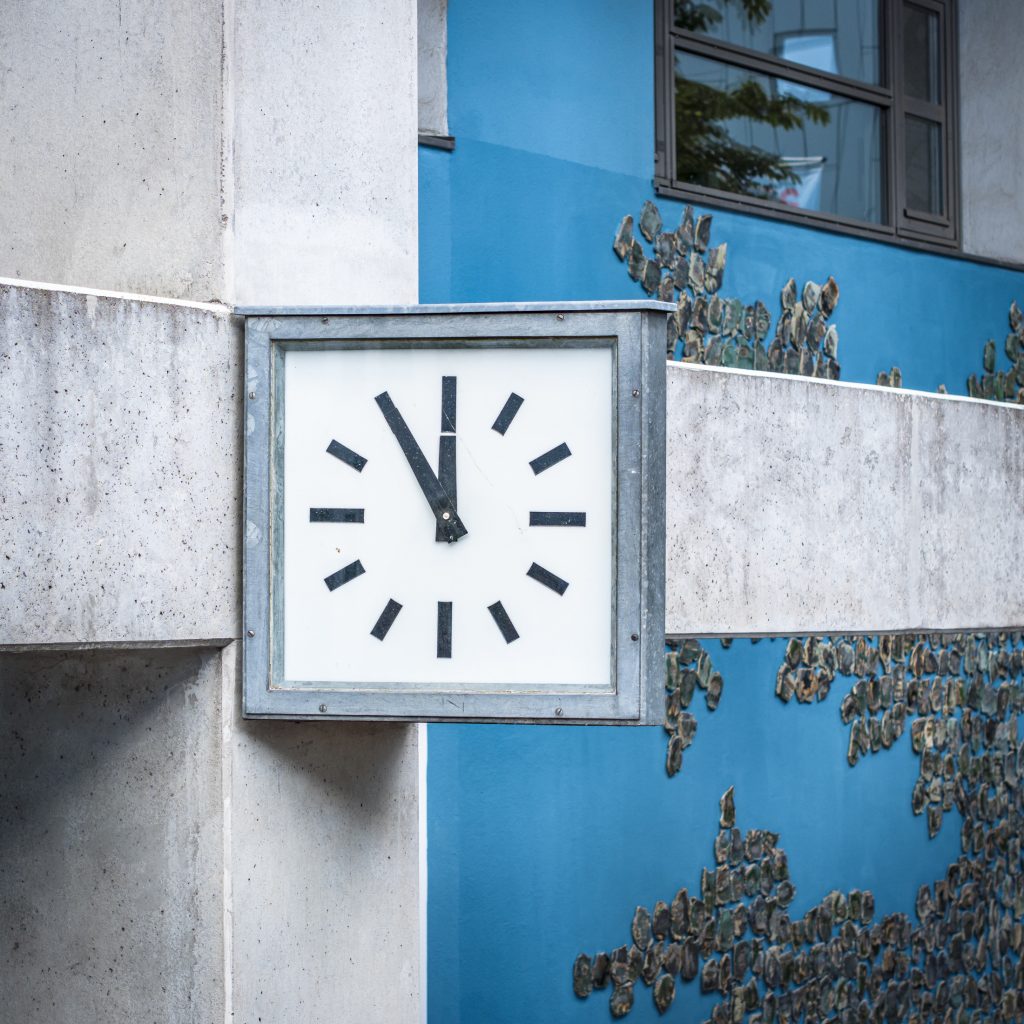 Uhr an Betonträger vor blauer Wand mit Kunstinstallation, Zeiger stehen auf fünf vor zwölf.