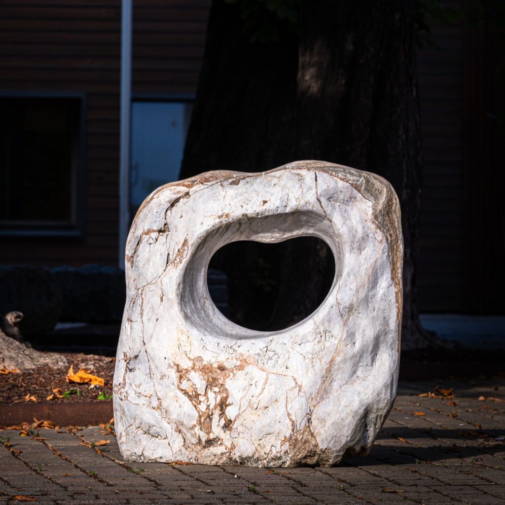 Heller Stein mit herzförmiger Öffnung, auf Pflaster vor Bäumen und Gebäudestruktur platziert.