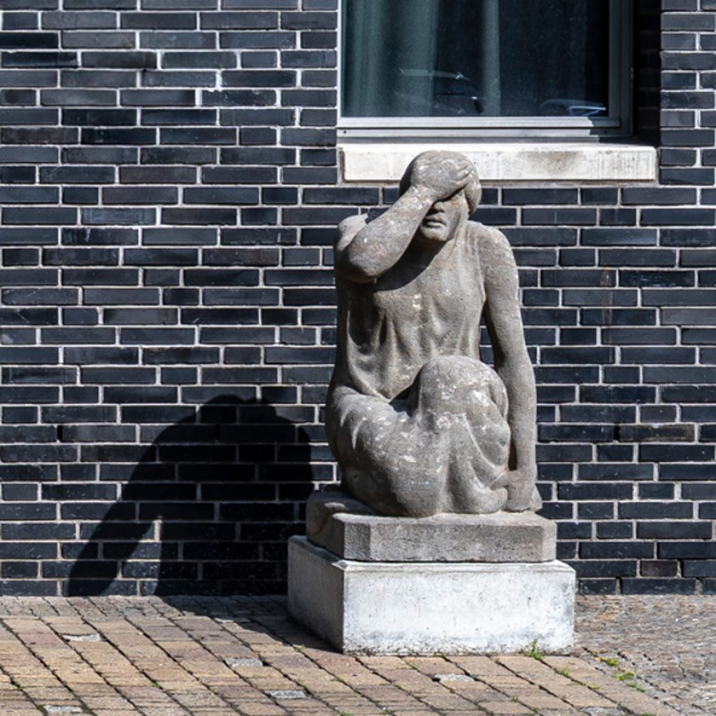 Skulptur einer sitzenden Figur mit erhobener Hand vor dem Gesicht, platziert vor dunkler Backsteinwand.