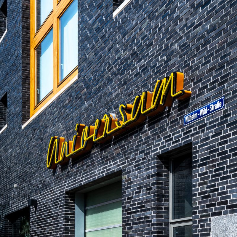 Gelber Schriftzug „Universum“ an dunkler Backsteinwand mit Straßenschild Wilhelm-Wild-Straße.