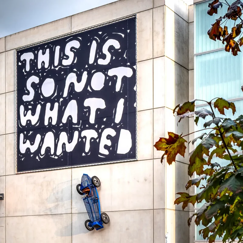 Großes schwarz-weißes Banner von Stefan Marx mit Text und Sportwagen-Skulptur an Betonfassade.